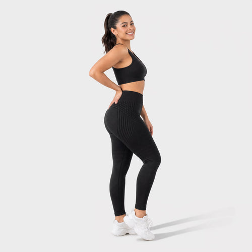 A woman modeling a matching black activewear set consisting of a sports bra and high-waisted ribbed leggings, paired with white sneakers, against a light gray studio background.