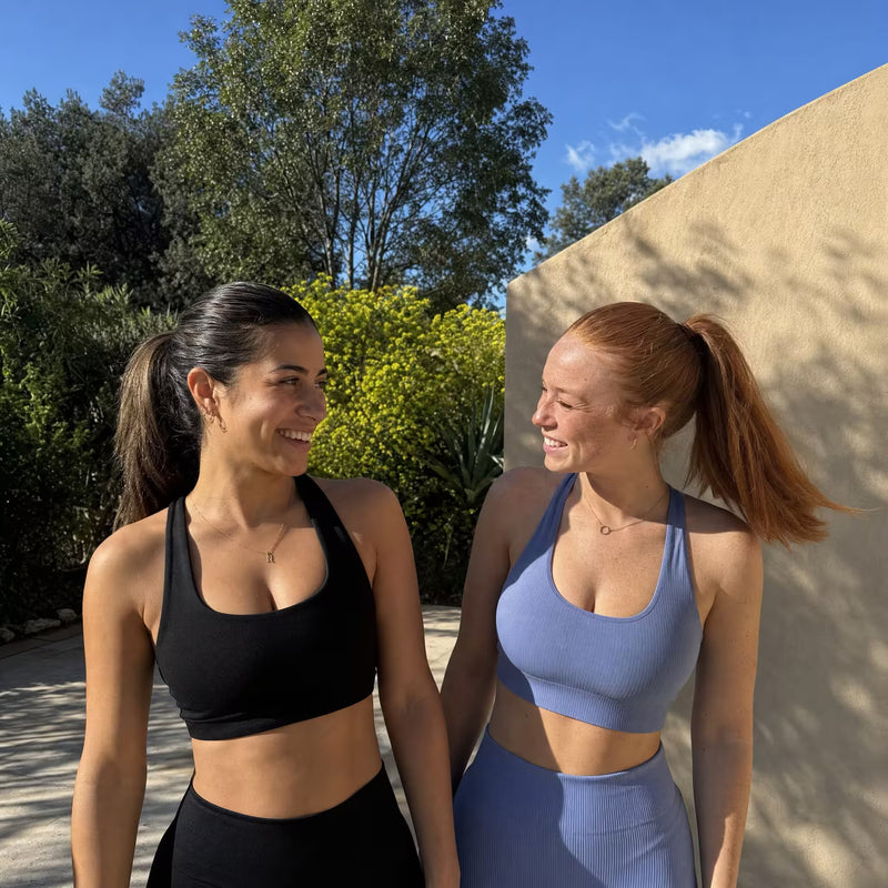 Two women standing outdoors next to each other, smiling and looking at one another. The woman on the left wears a black sports bra and black bottoms, and the woman on the right wears a dusty blue sports bra and matching blue bottoms.