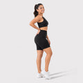 A woman modeling a black ribbed sports bra and matching high-waisted black compression shorts against a light gray studio background.