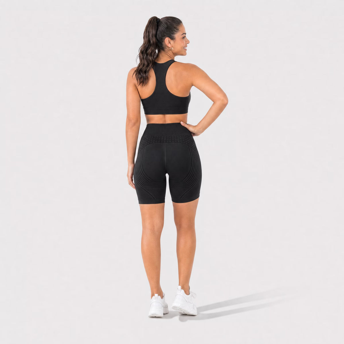 A woman is shown from the back wearing a matching black activewear set consisting of a sports bra and high-waisted biker shorts, paired with white sneakers, against a light gray studio background.