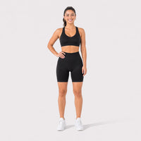 A woman modeling a black sports bra and matching high-waisted black biker shorts with white sneakers, standing against a light gray studio background.