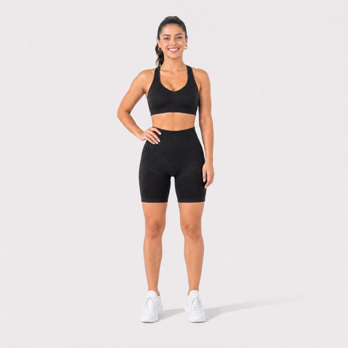 A woman modeling a black sports bra and matching high-waisted black biker shorts with white sneakers, standing against a light gray studio background.